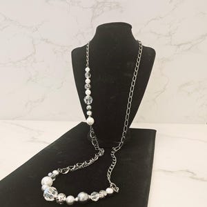 Puede incluir: Un collar largo de cadena plateada con una hebra de cuentas blancas y transparentes. El collar se muestra en un soporte de terciopelo negro.