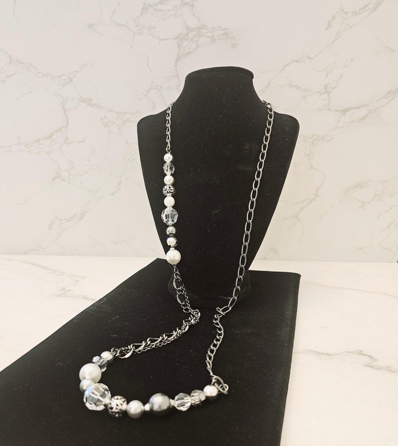 Puede incluir: Un collar largo de cadena plateada con una hebra de cuentas blancas y transparentes. El collar se muestra en un soporte de terciopelo negro.