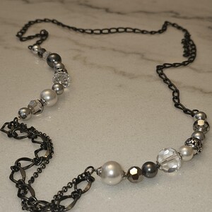 Puede incluir: Un collar de cadena plateado y negro con una variedad de cuentas plateadas y blancas. El collar presenta una mezcla de cuentas redondas, facetadas y con forma de perla.