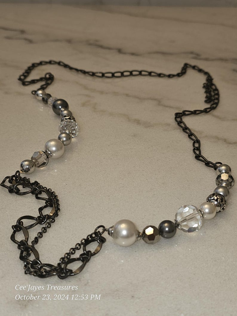 Puede incluir: Un collar de cadena plateado y negro con una variedad de cuentas plateadas y blancas. El collar presenta una mezcla de cuentas redondas, facetadas y con forma de perla.