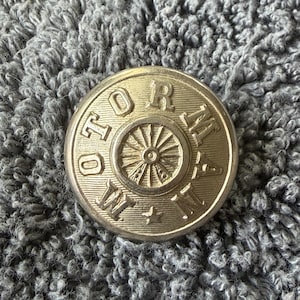 May include: A circular gold-toned metal button featuring a raised wheel design in the center. The word MOTORMAN is embossed in capital letters around the perimeter, with a small five-pointed star positioned at the bottom between the letters.