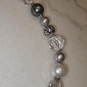Puede incluir: Un collar de cuentas plateadas y grises con cuentas de cristal transparente y una cuenta plateada con un dise&ntilde;o decorativo. El collar est&aacute; sobre una superficie blanca.