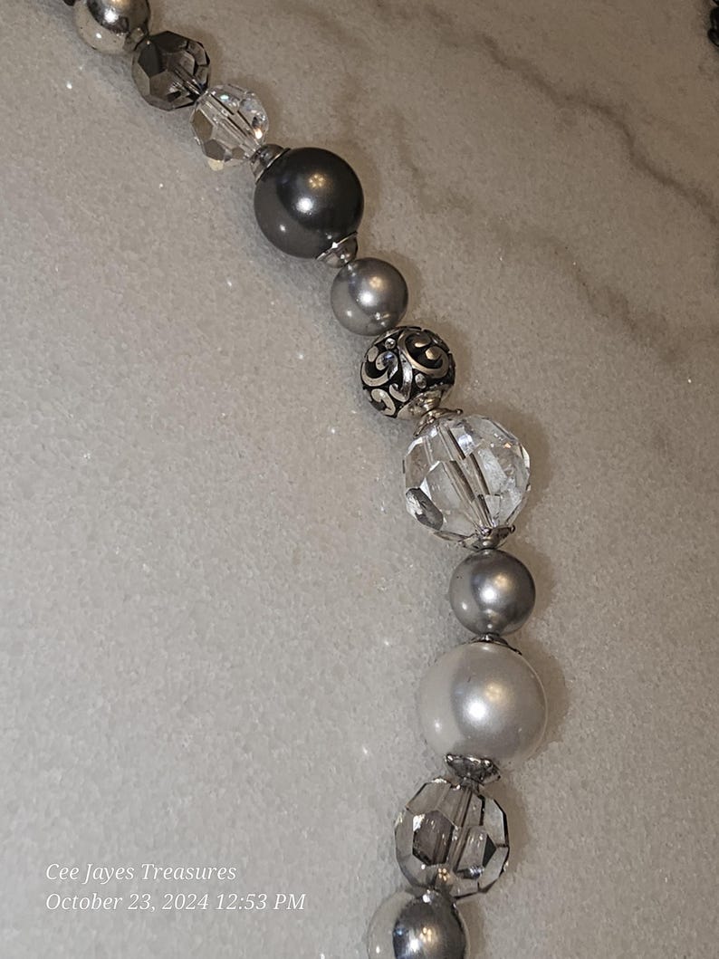 Puede incluir: Un collar de cuentas plateadas y grises con cuentas de cristal transparente y una cuenta plateada con un dise&ntilde;o decorativo. El collar est&aacute; sobre una superficie blanca.
