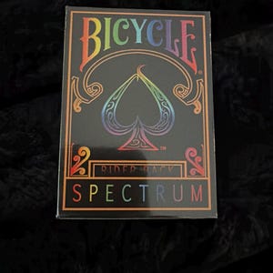 May include: A Bicycle Spectrum playing card deck. The black card box features a rainbow spade design and the word "Bicycle" in rainbow colors. The words "Rider Back" and "Spectrum" are also displayed on the box.