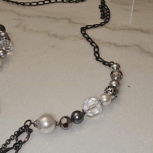 Puede incluir: Un collar de cuentas plateadas y negras con una cadena. Las cuentas son una mezcla de transparentes, blancas y grises.