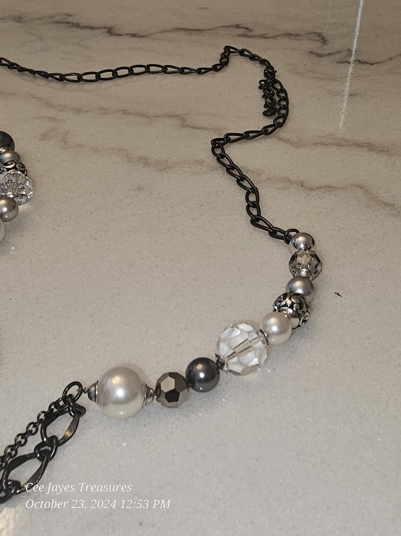 Puede incluir: Un collar de cuentas plateadas y negras con una cadena. Las cuentas son una mezcla de transparentes, blancas y grises.