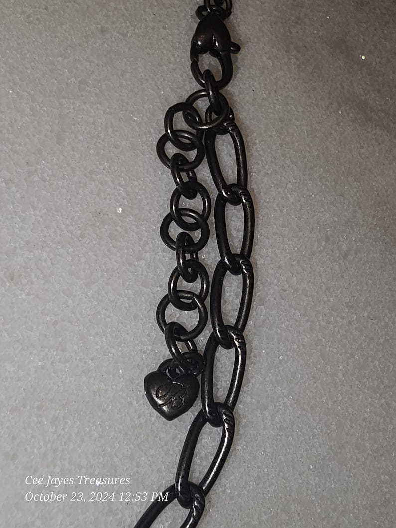 Puede incluir: Un collar de cadena de metal negro con un cierre en forma de coraz&oacute;n. El cierre tiene un peque&ntilde;o dije en forma de coraz&oacute;n con la letra "B" en &eacute;l.
