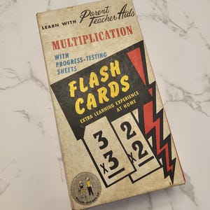Puede incluir: Una caja vintage de tarjetas de memoria de multiplicación con hojas de prueba de progreso. La caja es amarilla con acentos rojos y negros y presenta un gráfico de rayo. El texto en la caja dice "Aprende con Parent Teacher Aids Multiplicación con hojas de prueba de progreso Flash Cards Experiencia de aprendizaje adicional en casa".