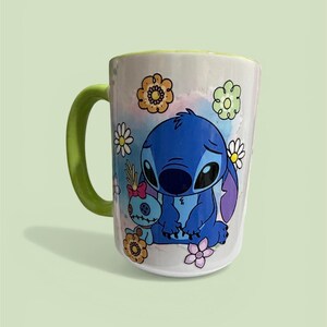 Pu&ograve; includere: Tazza in ceramica bianca con manico e bordo verdi, raffigurante un personaggio blu dei cartoni animati e un altro pi&ugrave; piccolo. La tazza &egrave; decorata con fiori colorati e uno sfondo ad acquerello. Un design divertente e stravagante.