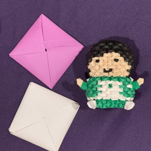 May include: A green and white origami figure of a person with black hair. The figure is wearing a green shirt and white pants. The figure is sitting on a purple surface with two folded paper squares, one pink and one white.