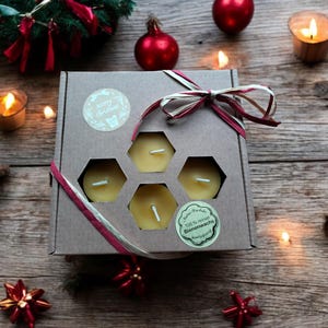 Könnte beinhalten: Eine Geschenkbox gefüllt mit sechs kleinen, runden, gelben Bienenwachskerzen. Die Box ist mit einem roten und weißen Band und einem "Frohe Weihnachten" Aufkleber verziert. Die Kerzen sind in einem Wabenmuster angeordnet.