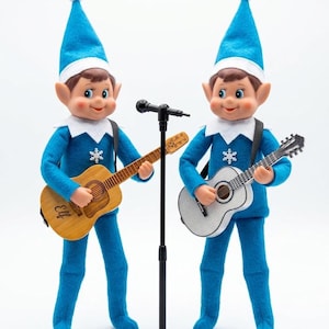 May include: Two elf dolls wearing blue felt outfits with white snowflake accents and matching blue hats with white pom-poms. One elf holds a light brown wooden guitar with the word Elf printed on it, while the other holds a silver glitter guitar.