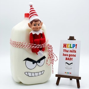 May include: A plastic milk jug with a cartoon face, wrapped in red and white twine, featuring a seated elf doll wearing a red and white striped hat. Next to it is a small wooden easel holding a sign that reads: HELP! The milk has gone BAD!