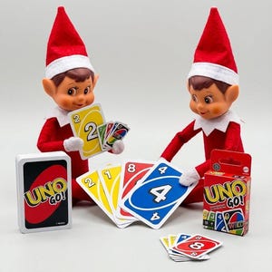 May include: Two elf dolls wearing red hats and outfits are playing a card game. One elf holds a yellow two card, while the other places a blue four card on a pile. The scene includes a deck of Uno Go! cards, a red Uno Go! box, and several scattered game cards.