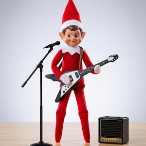 May include: A red elf doll wearing a pointed hat and white collar, standing next to a black microphone stand. The elf holds a black and white V-shaped electric guitar with a brown strap, and a small black amplifier sits on the floor nearby.