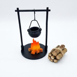 May include: A miniature black metal tripod stand with a hanging black cauldron positioned over a small orange and yellow flame figurine. Beside the stand is a small bundle of brown wooden sticks tied together with a thin piece of twine.