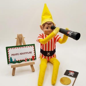 May include: A doll wearing a yellow hat, yellow sleeves, and a red and white striped shirt with a black belt featuring a skull emblem. The doll holds a black telescope with a skull and crossbones print. A wooden easel displays a sign reading PIRATE ADVENTURE, accompanied by a small black pirate flag and a gold coin.