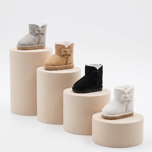 May include: Four ankle boots in gray, tan, black, and white, each featuring a small bow detail on the side, a rounded toe, and a thick sole. The boots are lined with a light-colored material and have a soft, textured exterior finish.