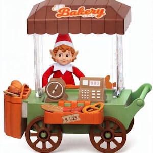 May include: A toy bakery cart with a brown roof labeled Bakery Shop, featuring a small elf figure in a red outfit. The green cart includes a brown scale, a cash register with 000 displayed, and a basket of bread with a $1.25 price tag.