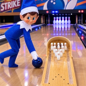 May include: A small elf figurine wearing a blue felt suit and matching pointed hat with a white collar and cuffs, positioned on a bowling lane. The elf is touching a small blue bowling ball in front of a miniature wooden bowling alley set.