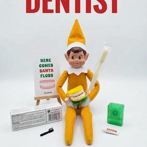 May include: A yellow elf doll holding a set of plastic teeth and a toothbrush in a clear wrapper. Beside it are a small sign reading "HERE COMES SANTA FLOSS", a box of toothpaste, a tiny black brush, a green dental floss container, and a small tube.
