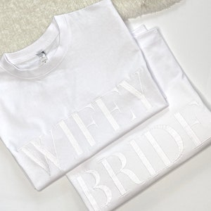 Könnte beinhalten: Zwei weiße T-Shirts mit den Worten "WIFEY" und "BRIDE" in weiß auf der Vorderseite gestickt.