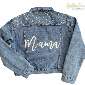 K&ouml;nnte beinhalten: Eine hellblaue Jeansjacke mit wei&szlig;en Perlenbesatz an den Schultern und dem Wort "Mama" in wei&szlig;er Stickerei auf der R&uuml;ckseite.