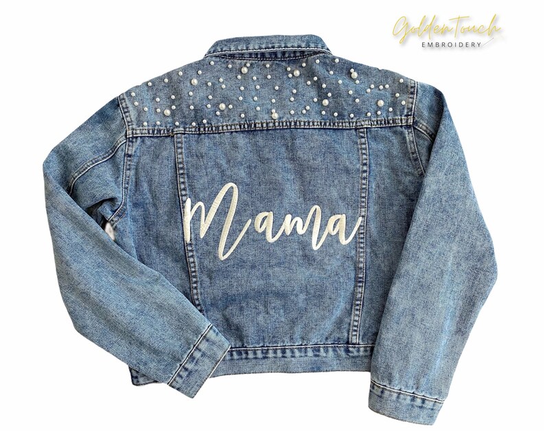 K&ouml;nnte beinhalten: Eine hellblaue Jeansjacke mit wei&szlig;en Perlenbesatz an den Schultern und dem Wort "Mama" in wei&szlig;er Stickerei auf der R&uuml;ckseite.