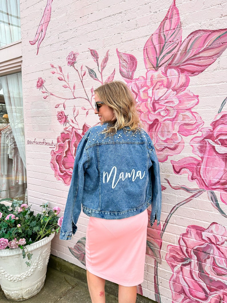 K&ouml;nnte beinhalten: Eine Frau tr&auml;gt eine Jeansjacke mit dem Wort "Mama" in wei&szlig; auf dem R&uuml;cken. Die Jacke ist mit Strasssteinen verziert. Sie steht vor einer pinkfarbenen Wand mit einem floralen Wandbild.