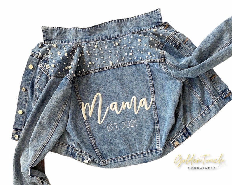 K&ouml;nnte beinhalten: Eine blaue Jeansjacke mit Perlenbesatz an den Schultern und dem aufgen&auml;hten Wort "Mama" auf der Vorderseite, darunter der Text "EST. 2021".