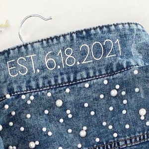 K&ouml;nnte beinhalten: Eine blaue Jeansjacke mit wei&szlig;en Perlenbesatz und wei&szlig;er Stickerei mit der Aufschrift "EST. 6.18.2021".