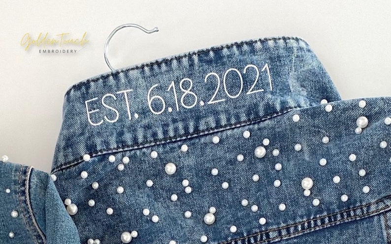 K&ouml;nnte beinhalten: Eine blaue Jeansjacke mit wei&szlig;en Perlenbesatz und wei&szlig;er Stickerei mit der Aufschrift "EST. 6.18.2021".