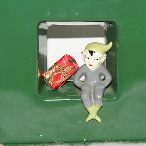 May include: A green ceramic Christmas tree with a small elf figurine holding a wrapped gift in a window opening.