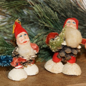 May include: Two vintage Christmas ornaments of Santa Claus figures. The figures have red hats and coats, white beards, and are made from pine cones. One holds a blue tree, the other a green garland. The ornaments are on a wooden surface.