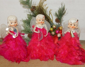 May include: Three vintage Christmas angel tree toppers with pink feather skirts and clear plastic wings. The angels are holding musical instruments and are made of porcelain.