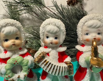 Op de afbeelding: Drie vintage porseleinen kerstfiguurtjes van kinderen die rode jassen met witte bontkragen en witte hoeden dragen. De kinderen spelen muziekinstrumenten: een meisje met een groene krans, een meisje met een accordeon en een meisje met een gouden saxofoon.
