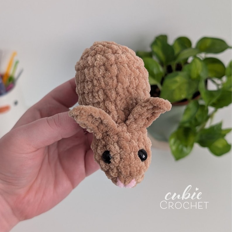 Op de afbeelding: Een handgemaakt, lichtbruin gehaakt konijn met zwarte ogen en een roze neus. Het konijn heeft lange oren en een zachte textuur. De tekst "cubie CROCHET" staat rechtsonder.