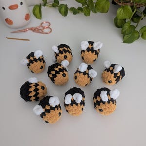 May include: A collection of handmade crochet bees in black, yellow, and white. The small, round bees have black and yellow striped bodies, white wings, and small black eyes. The bees are arranged on a white surface.