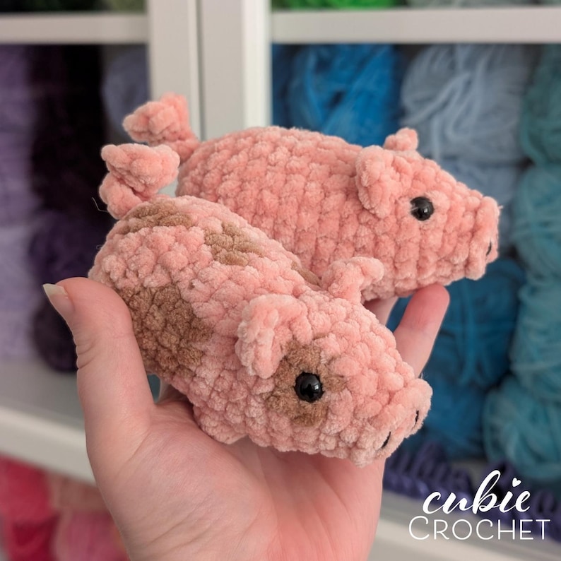 May include: Two small, handmade crochet pigs crafted from soft, light pink plush yarn. The pigs feature dark brown patches, small pointed ears, and black plastic eyes. One pig has a curled tail, and both have a snout with two small black dots.