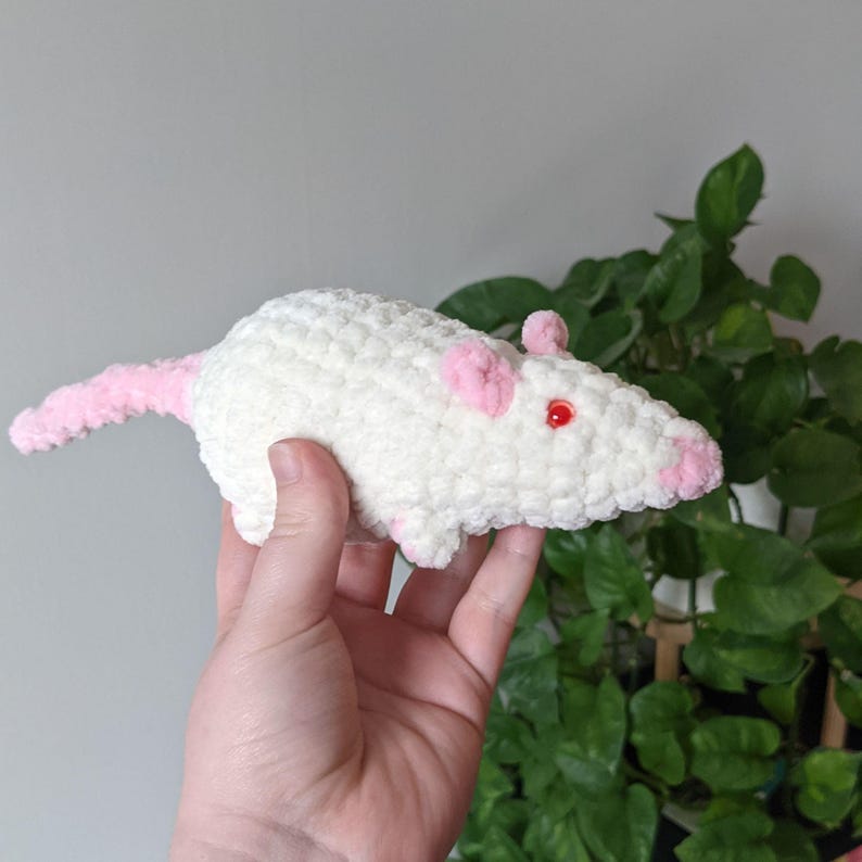 A cream colored crochet rat plushie with red eyes, light pink nose, ears, and tail, being held up by a hand in front of a pothos plant.