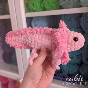 May include: A handmade pink crochet axolotl plush toy. The plush features a light pink body with darker pink frills and a small, black eye. The axolotl is held in a hand, with the "cubie CROCHET" logo in the bottom right corner.