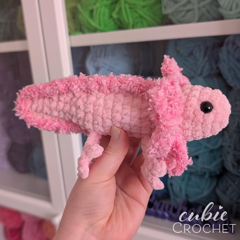 May include: A handmade pink crochet axolotl plush toy. The plush features a light pink body with darker pink frills and a small, black eye. The axolotl is held in a hand, with the "cubie CROCHET" logo in the bottom right corner.