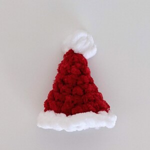 May include: A small, red Santa hat with a white, fluffy trim and a white pom-pom at the top. The hat is made of a textured, plush material, perfect for holiday decorations or festive wear.