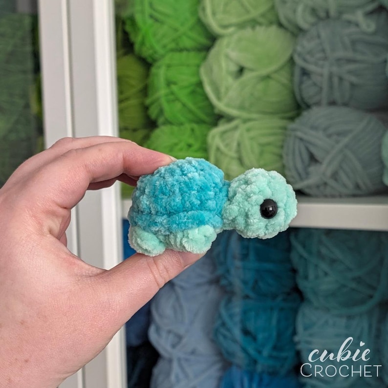 May include: A small, plush, turquoise-colored crocheted turtle toy with a soft, fuzzy texture. The turtle features a rounded shell, a small head with a single visible black plastic eye, and four short, stubby legs. The yarn has a velvet-like finish.