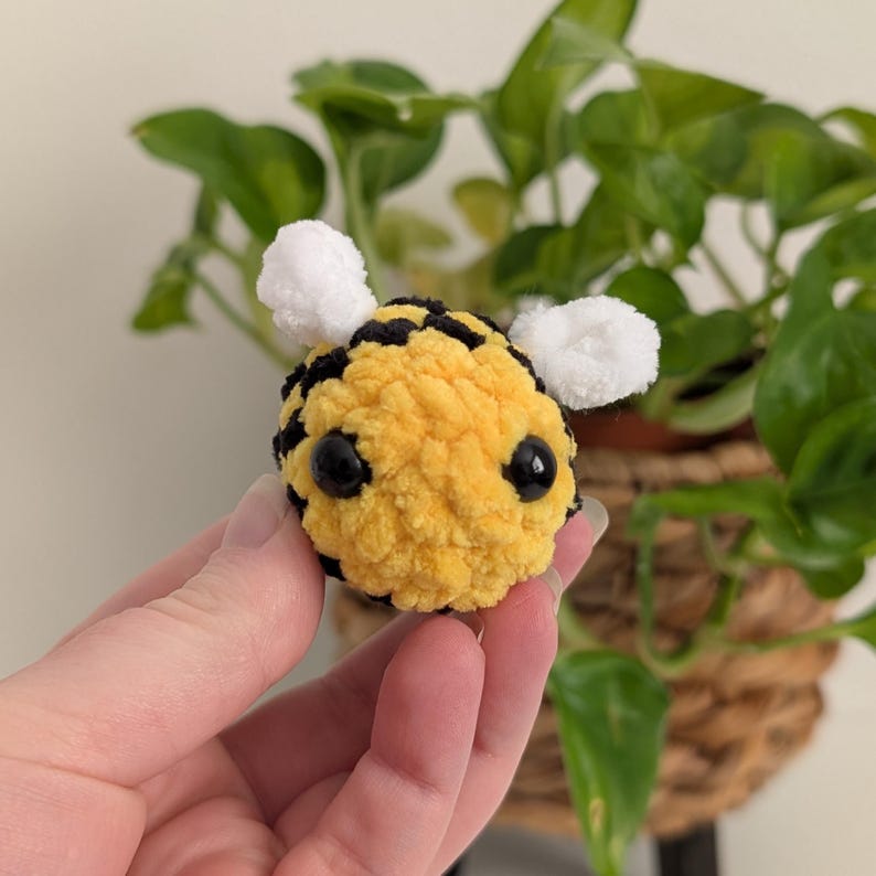 May include: A handmade, plush bee with a yellow body, black stripes, and white wings. The bee has large, black eyes and is made of soft, textured yarn. A cute, decorative item.