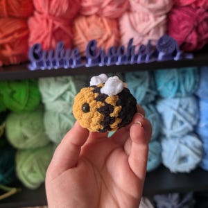 May include: A handmade crochet bee with yellow, black, and white accents. The bee has a round body, small black eyes, and fluffy white wings. The background features colorful yarn skeins and the text "Cubie Crochet".