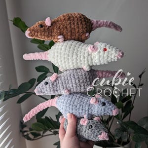 A stack of 5 crochet rat plushies, balanced in one hand. Each rat has light pink nose, ears, and tail. The bottom one is dark gray, the next two up are different shades of light gray, the fourth one up is cream-colored, and the top one is brown. The cream-colored one has red eyes, while the rest have black eyes.