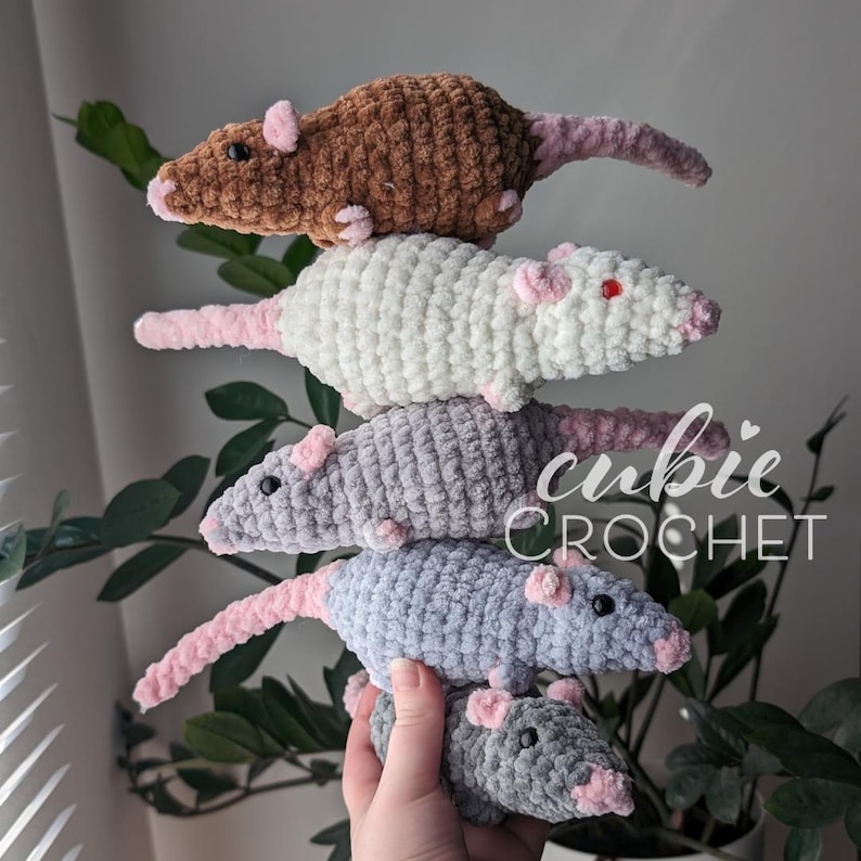 A stack of 5 crochet rat plushies, balanced in one hand. Each rat has light pink nose, ears, and tail. The bottom one is dark gray, the next two up are different shades of light gray, the fourth one up is cream-colored, and the top one is brown. The cream-colored one has red eyes, while the rest have black eyes.