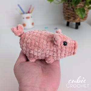 May include: A small, handmade crochet pig toy crafted from soft, light pink plush yarn. The pig features a rounded body, a curly tail, two small ears, and a black plastic bead eye. The texture is fuzzy and uniform throughout the entire figure.