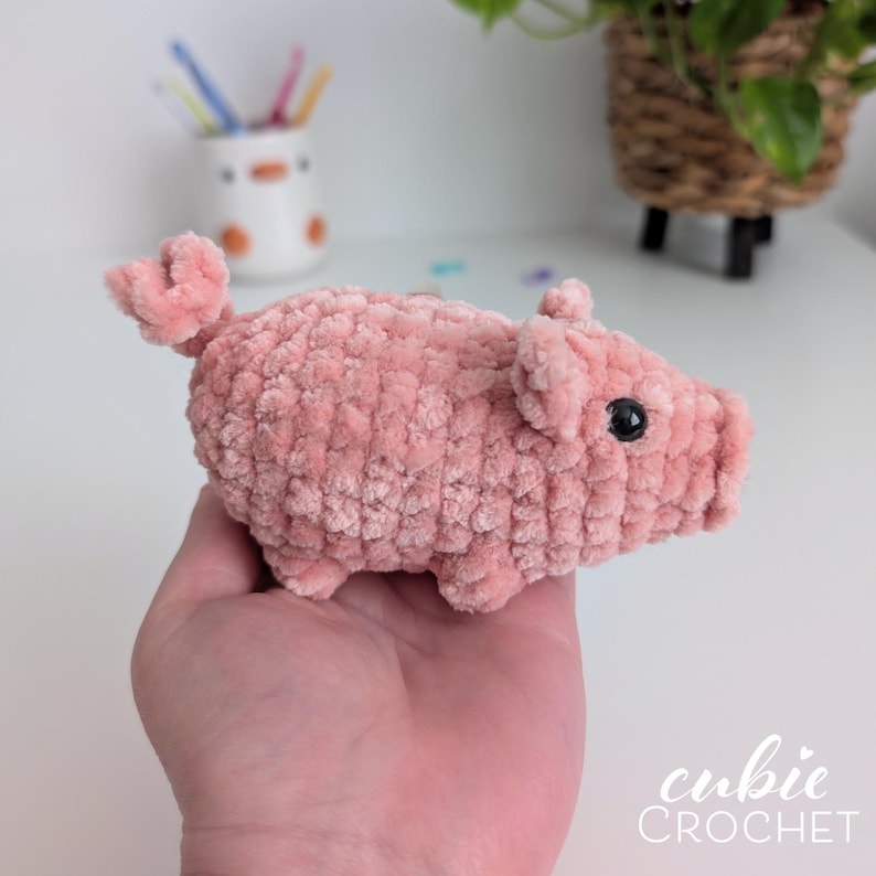 May include: A small, handmade crochet pig toy crafted from soft, light pink plush yarn. The pig features a rounded body, a curly tail, two small ears, and a black plastic bead eye. The texture is fuzzy and uniform throughout the entire figure.